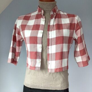 chemise crop top fond blanc, motif plaid rose. Photo sur mannequin. la chemise est présentée de face, ouverte sur un pull beige à col roulé et une jupe noire.